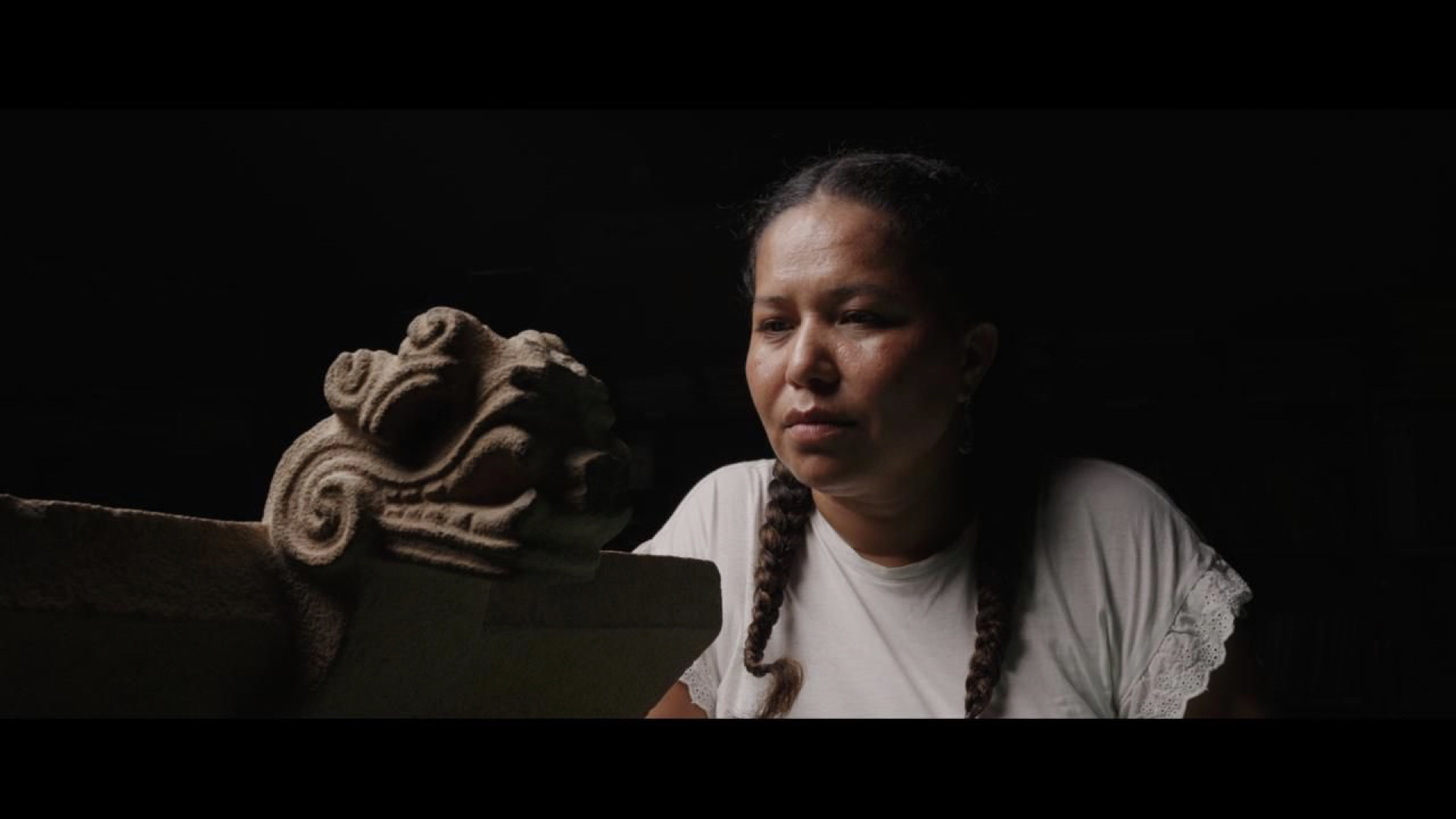 A woman with braided hair, wearing a light shirt, looks intently at a carved stone sculpture with a stylized face.