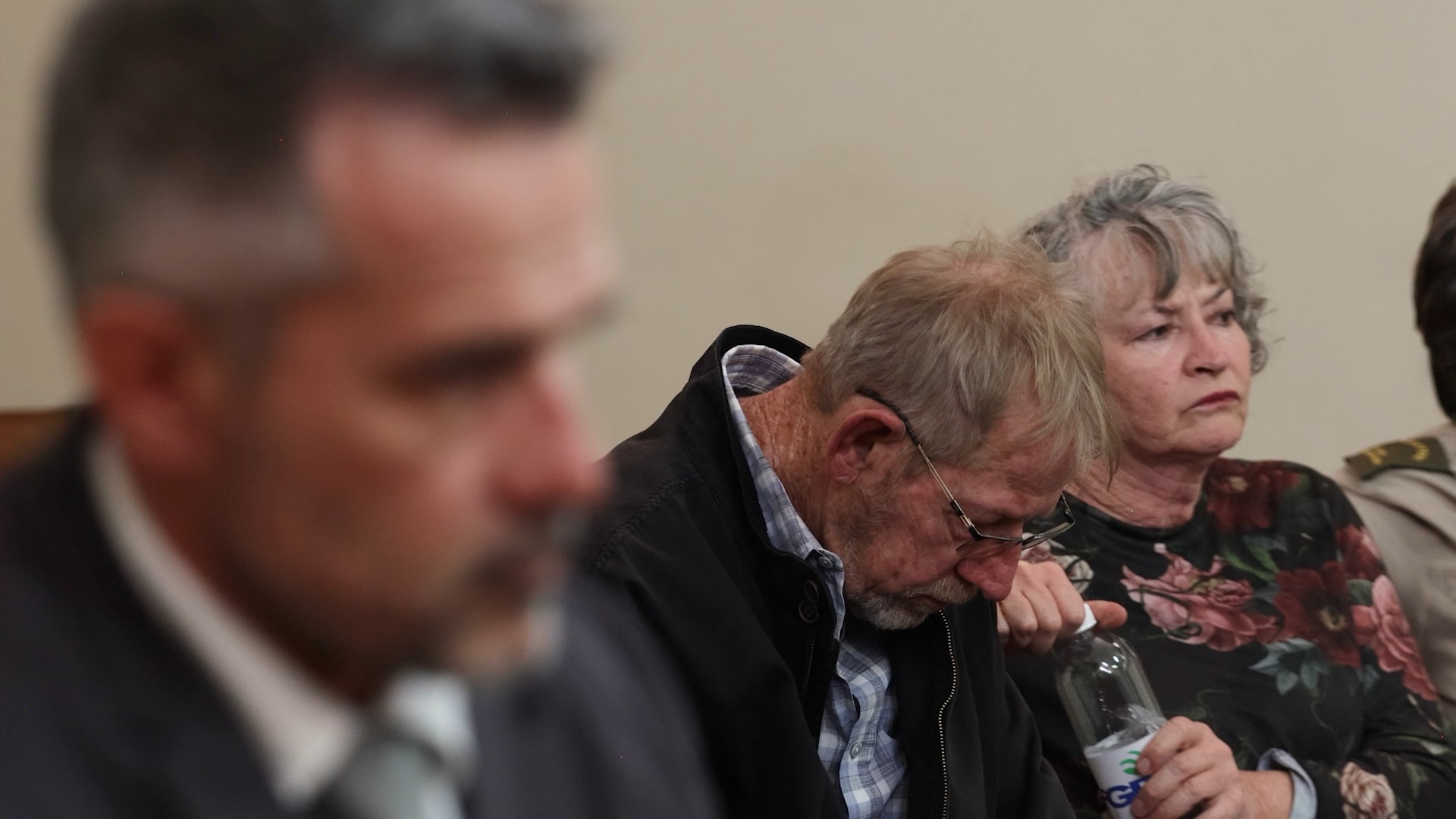 Three people seated indoors; a blurred man in front, while an older man looks down holding a bottle and an older woman looks ahead seriously.