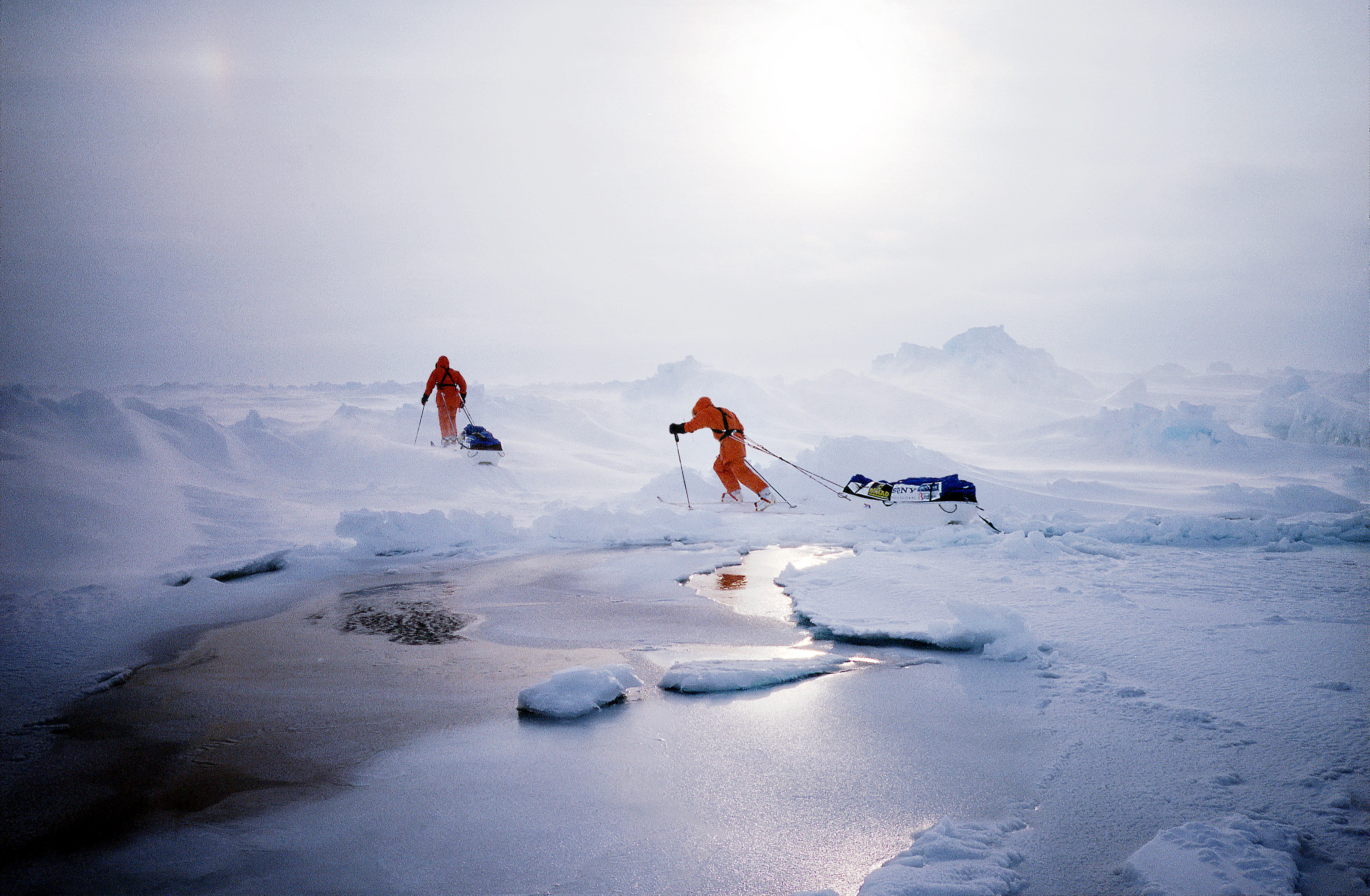 Two people in bright orange cold-weather suits ski across a vast, icy landscape in blowing snow, each pulling a sled behind them. 