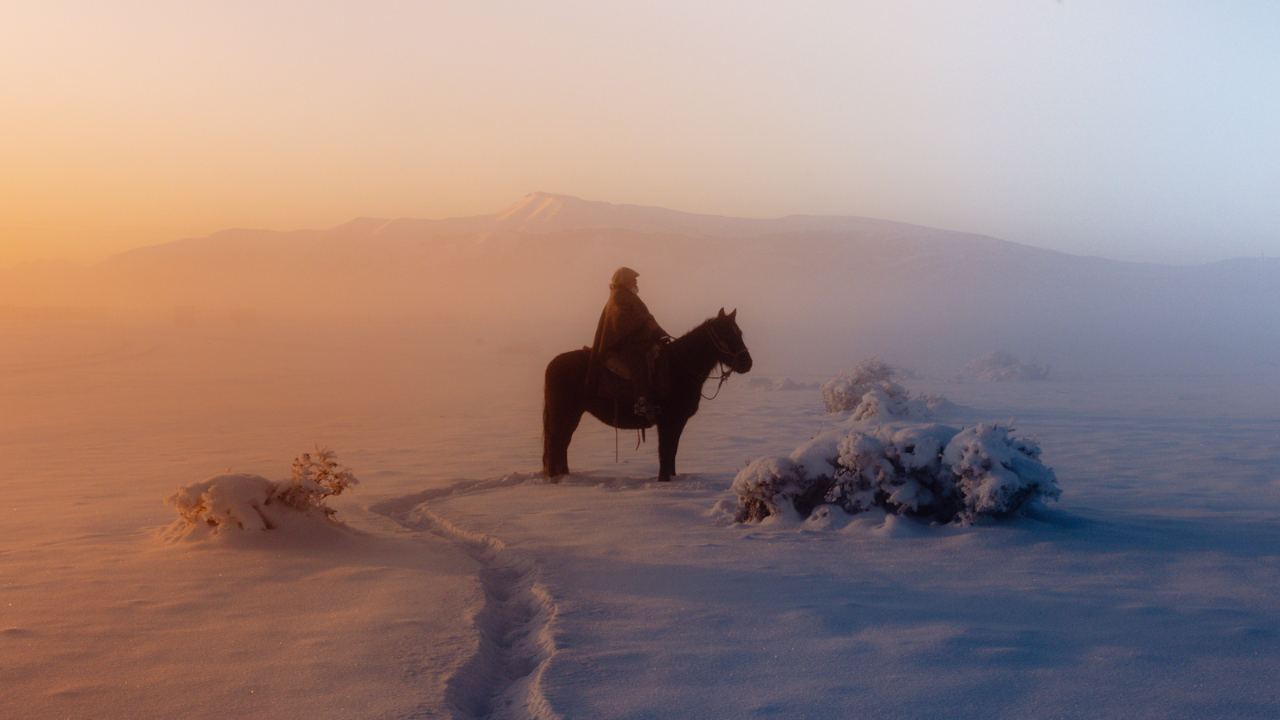 A silhouette of a man sat on a horse in the centre, in a snowy mountain range with a purple and orange haze coming from the left side. 