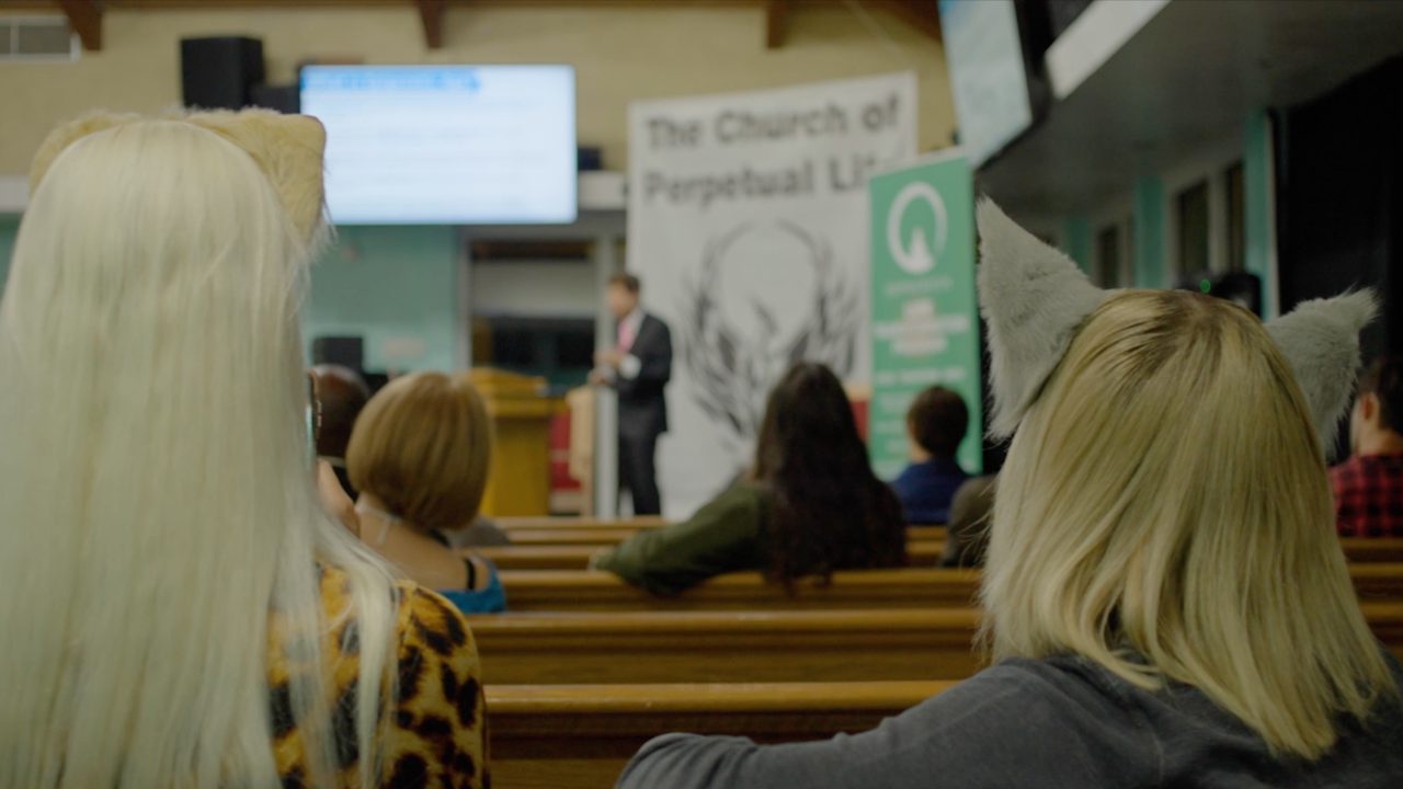 Two people wearing animal-ear headbands sit in church pews facing a speaker at the front of a sanctuary, with banners and a presentation screen in the background