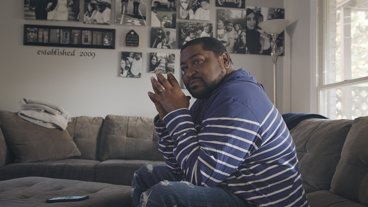 A black man in a striped shirt sits on a couch, hands clasped, looking thoughtfully toward the camera, with family photos on the wall behind him.