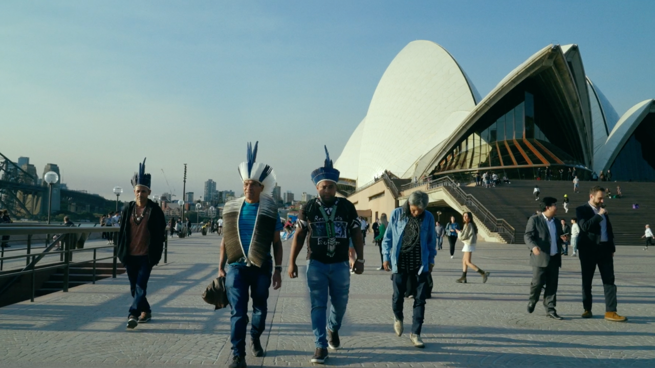Four Indigenous men wearing feathered ceremonial headdresses walk along the waterfront promenade in front of the Sydney Opera House, with other pedestrians nearby and the Sydney Harbour Bridge visible in the background.