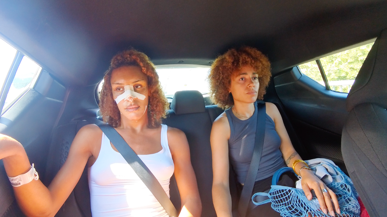 Two women sit in the backseat of a car wearing seatbelts. One has a bandaged nose and a hospital wristband, looking tired, while the other looks ahead quietly, holding a blue bag.