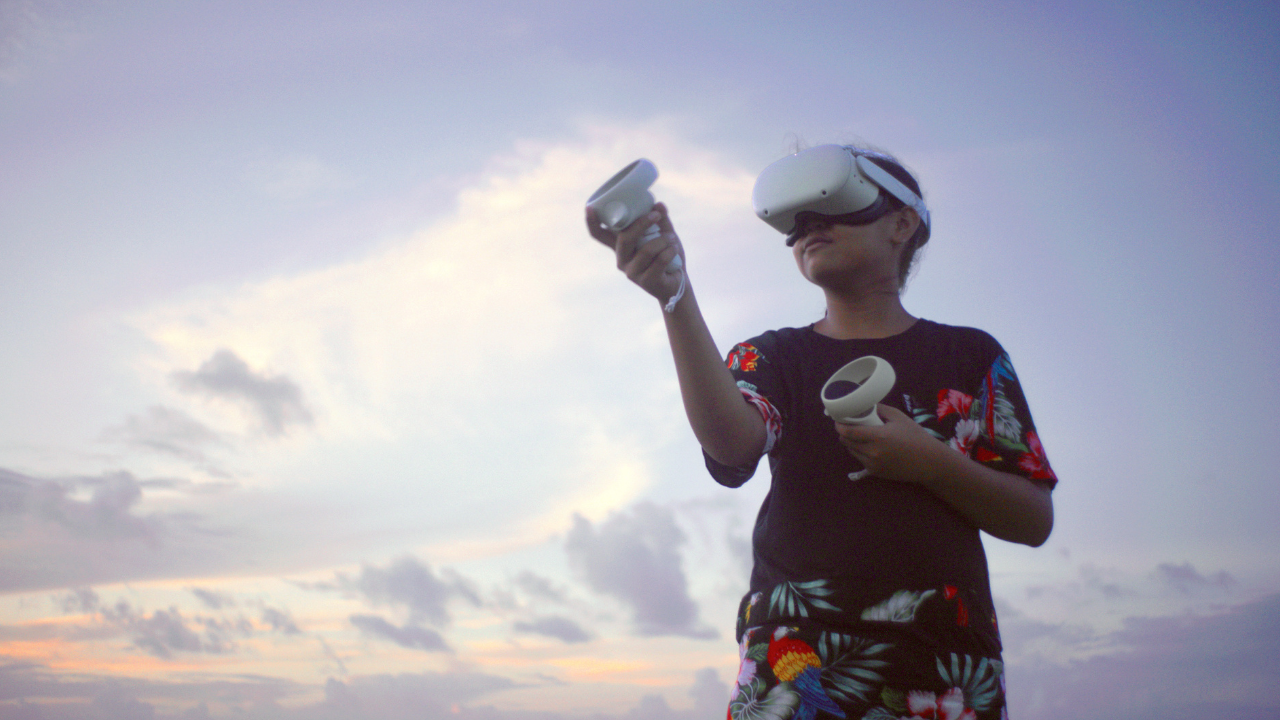 A child wearing a virtual reality headset stands outdoors under a cloudy sky at dusk, holding two motion controllers while playing or exploring in VR. The child wears a black shirt and shorts with colorful tropical floral patterns.
