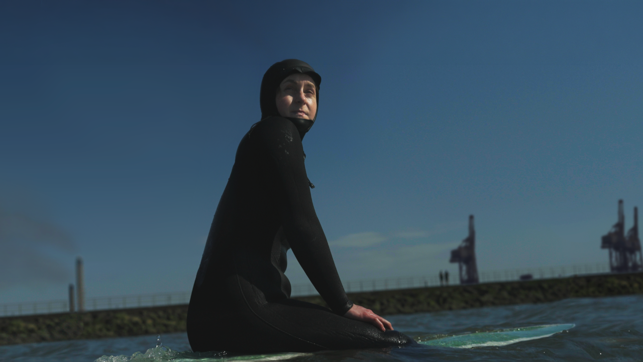 A person in a black wetsuit sits on a surfboard in the water, looking off into the distance under a clear blue sky, with an industrial-looking shoreline and cranes in the background.