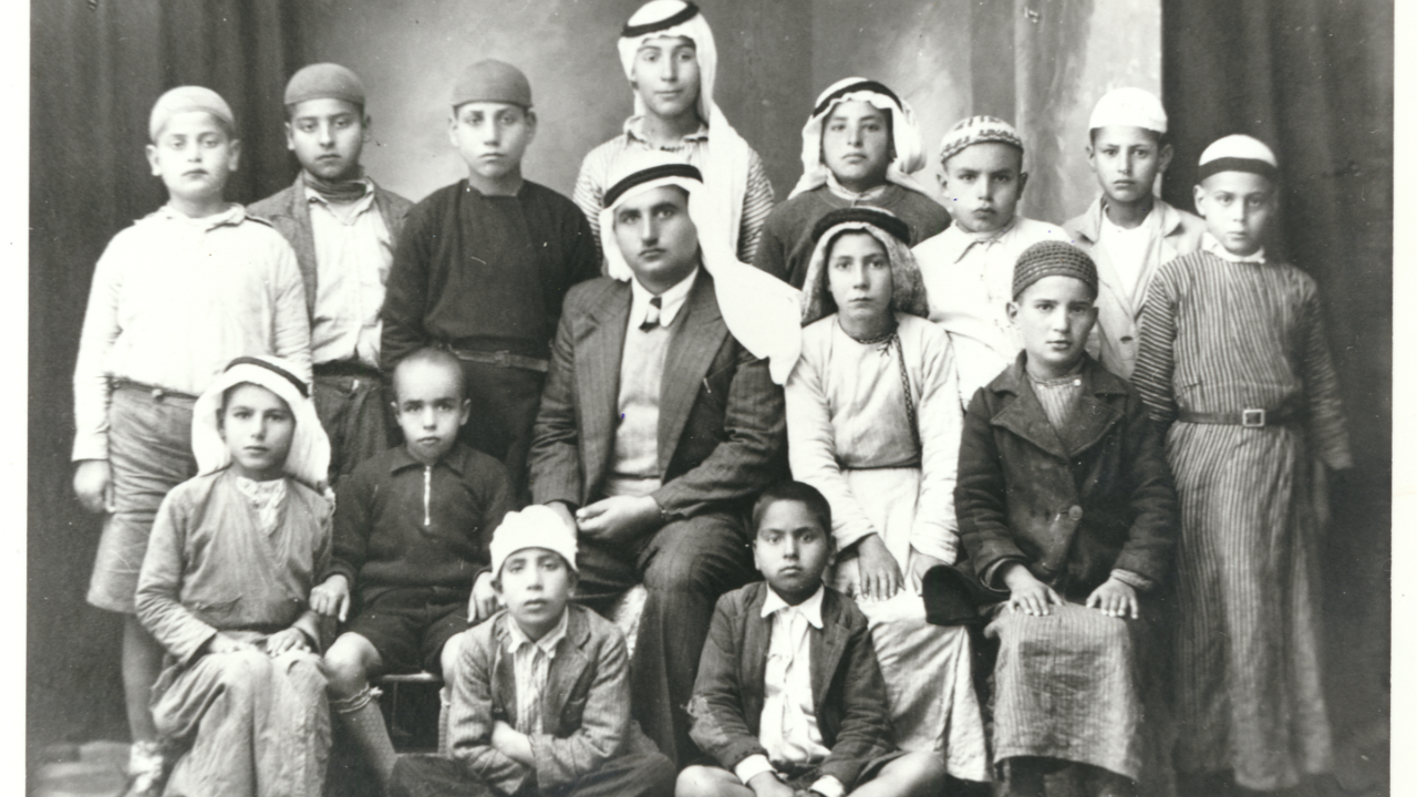A black and white image of a teacher sat in the centre wearing a keffiyeh, with his pupils surrounding. 