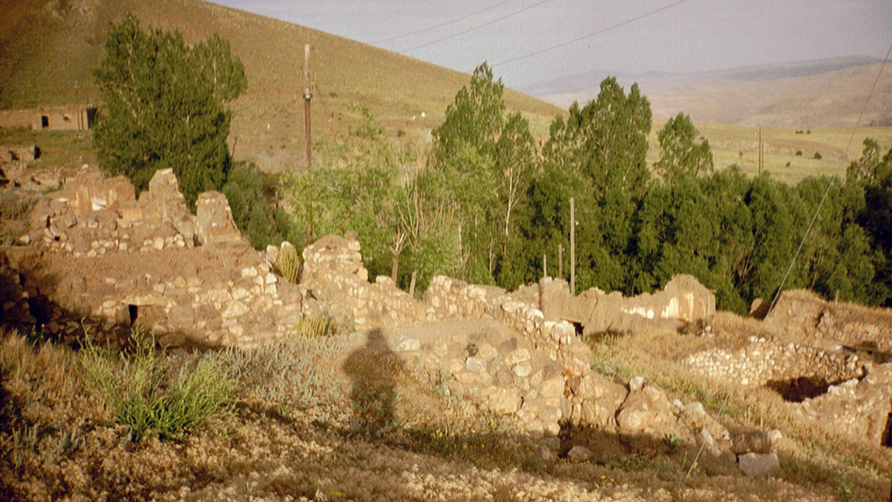 Rocky, ruined walls of a building have a shadow of a person cast over them, in the centre of the image. Behind them, a line of large green trees sits, with dusty, rolling hills in the background.