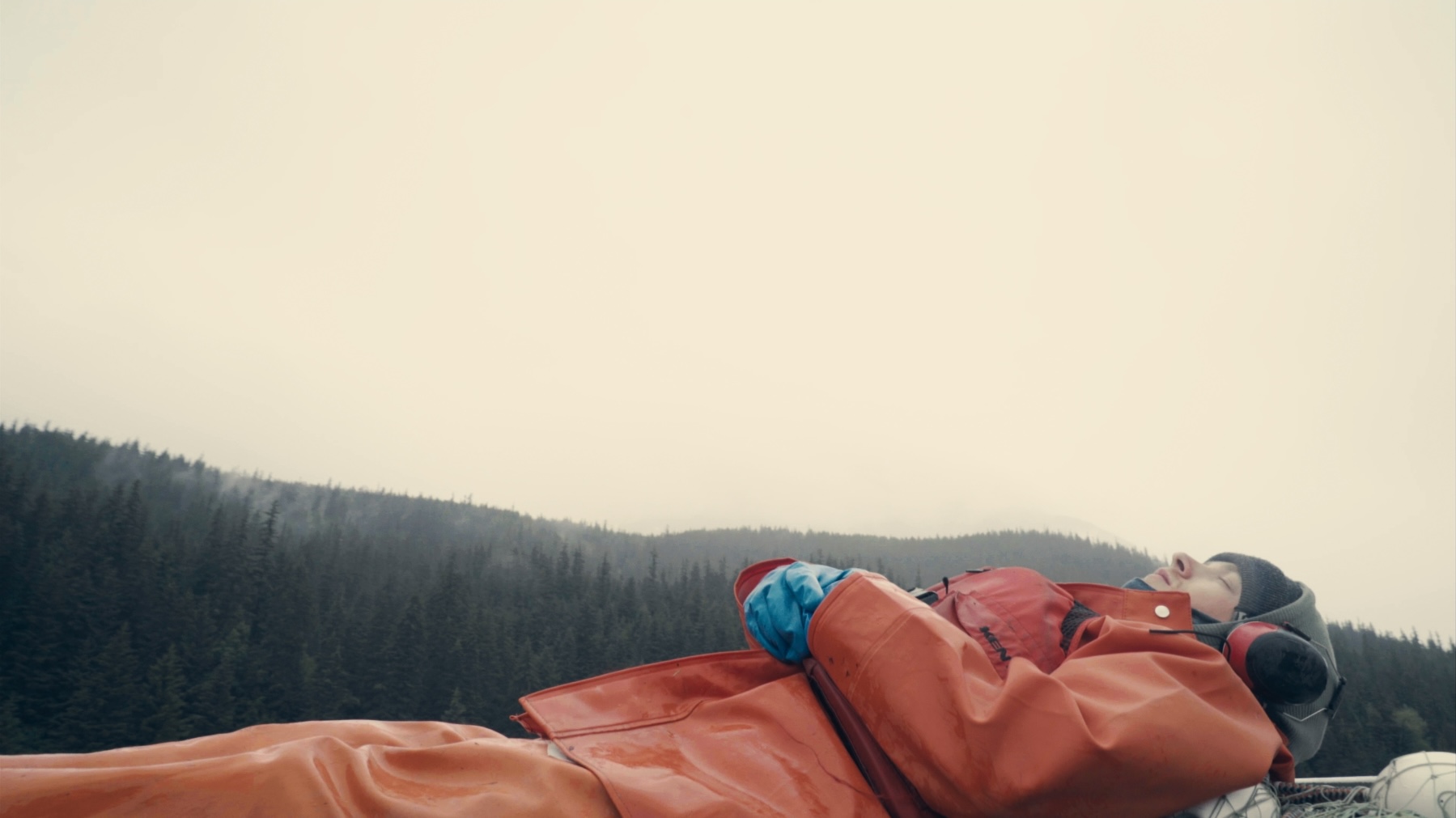 A person dressed in orange fisherman's clothes with red ear defenders on lays on his back in the bottom of the frame. In the background is a grey sky and a line of trees and mountains