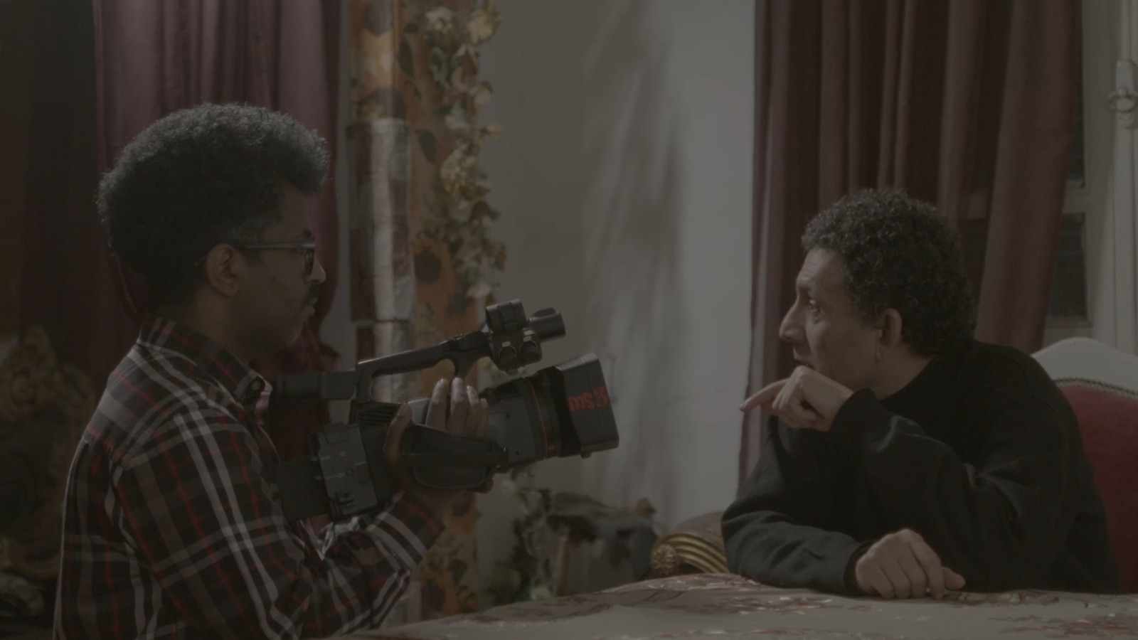 A man in a plaid shirt films another man seated across a table. The second man leans forward thoughtfully, creating an intimate, interview like indoor scene.