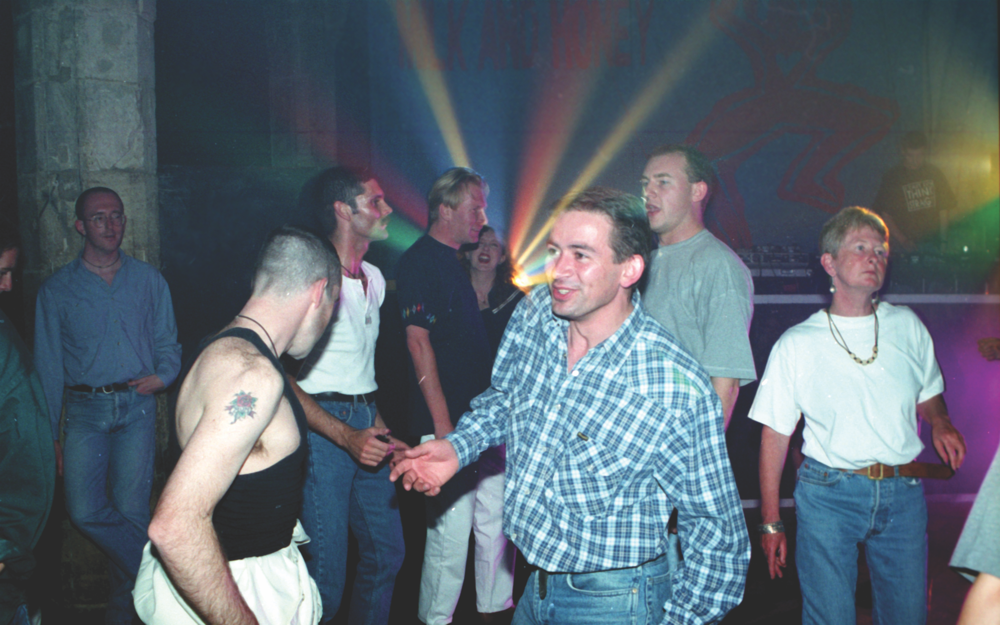 A group of people dance and socialize in a nightclub, with colorful lights shining behind them. Several individuals chat and move to the music, creating a lively, energetic atmosphere.