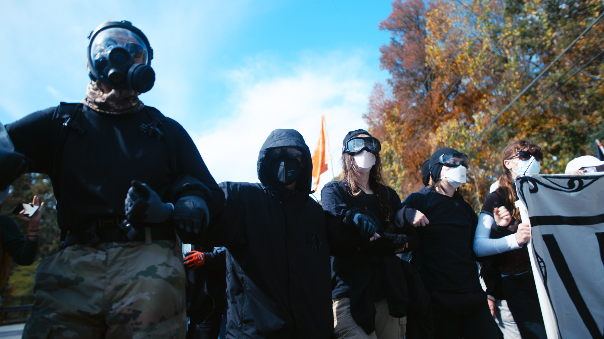 A group of activists are holding arms while wearing black outfits and face coverings to conceal their identities.
