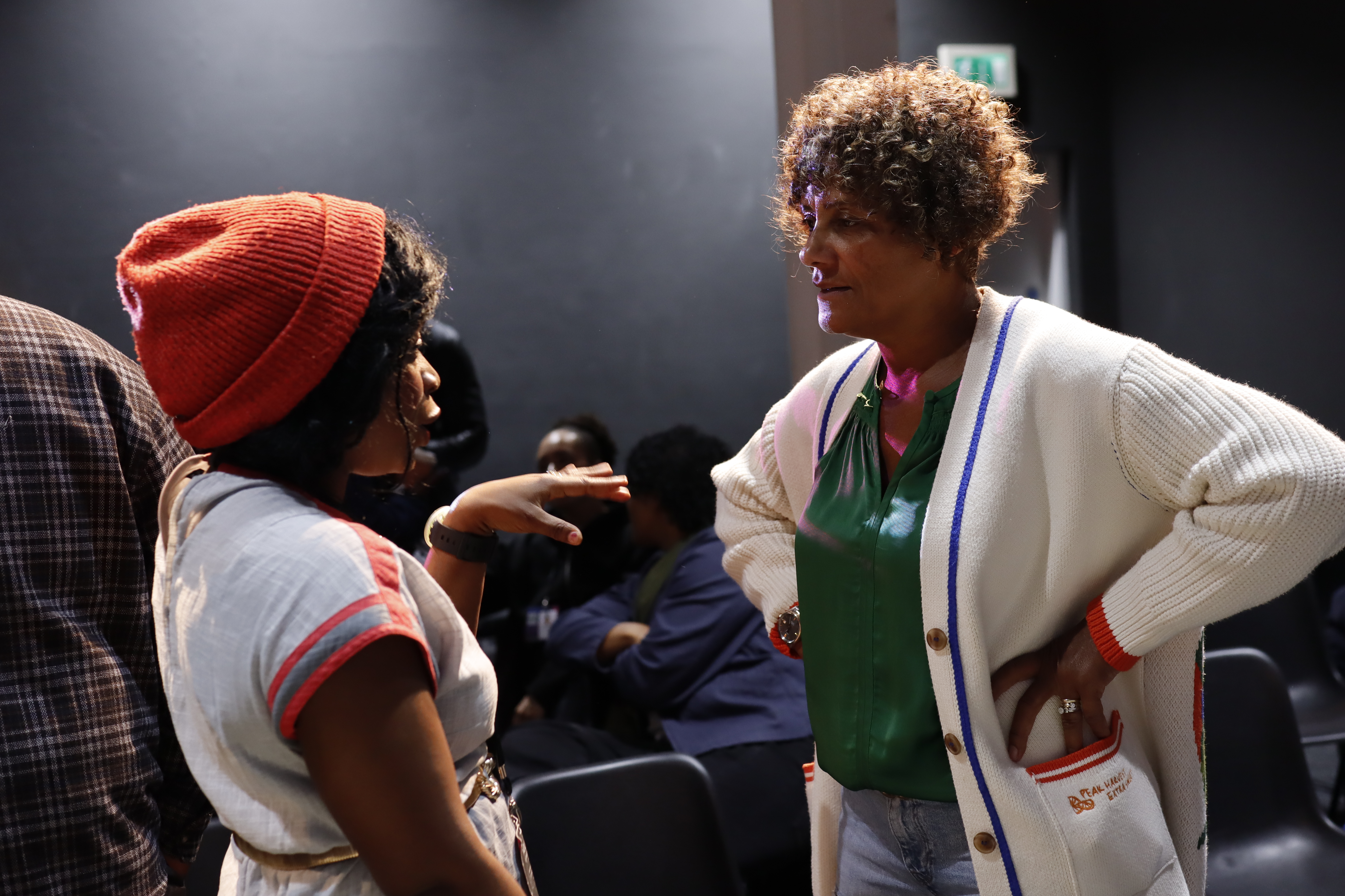 A woman with her hands on her hips talks to a woman in an orange hat.