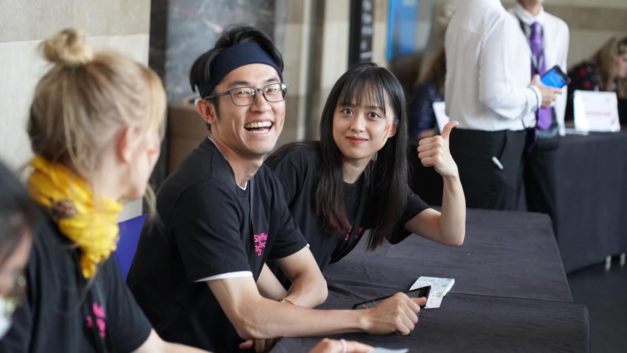 Three volunteers are sat at a long black table. Two face the camera smiling, one is gesturing a thumbs up to the camera