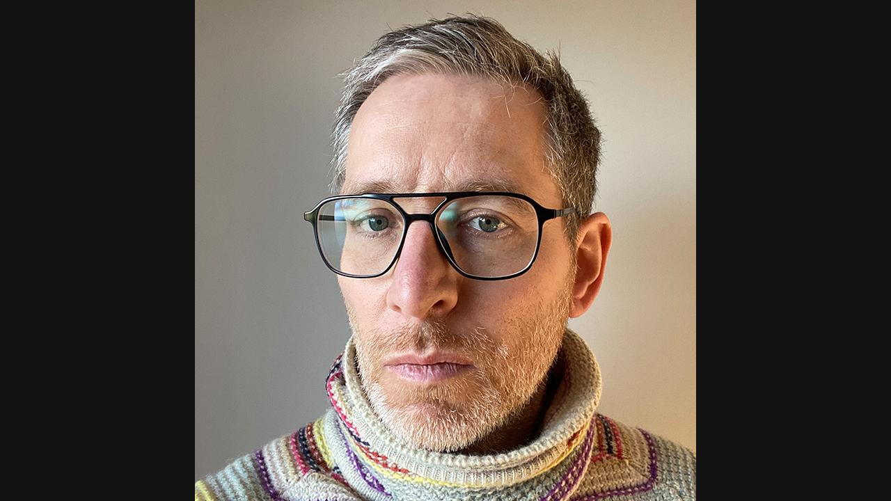 Mat Steel headshot. Image of a white man with square, black rimmed glasses. He's wearing a colourful knit jumper and is pictured against a neutral coloured wall.