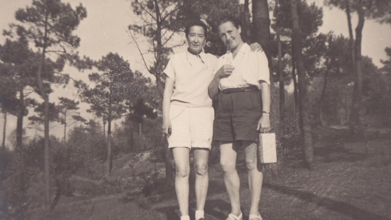 A sepia tone photograph of two women, wearing collared shirts and shorts. They're stood in a wooded area, in front of trees.