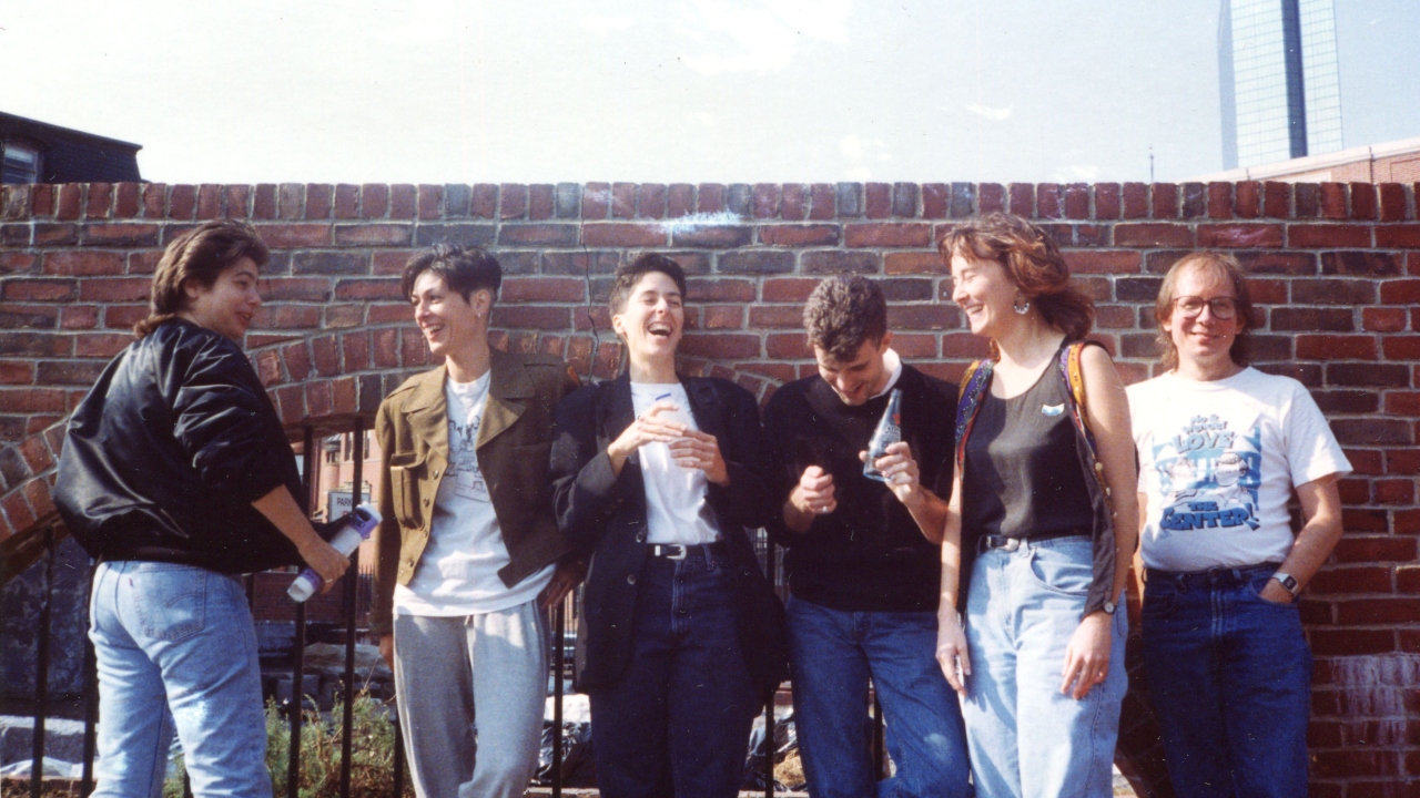 Archival image of a group of six people stood in a line beside a red brick wall. They are pictured smiling and laughing with each other.