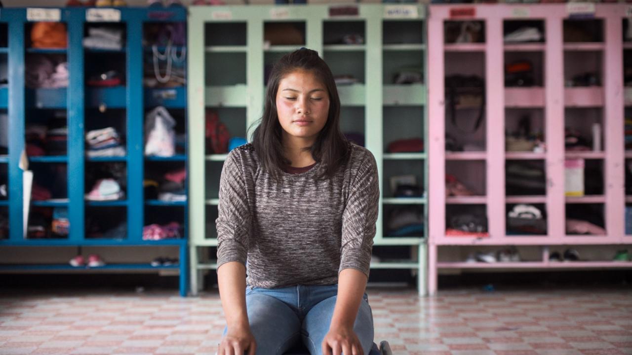 A young girl with long brown hair sits central to the camera, she has her eyes closed.