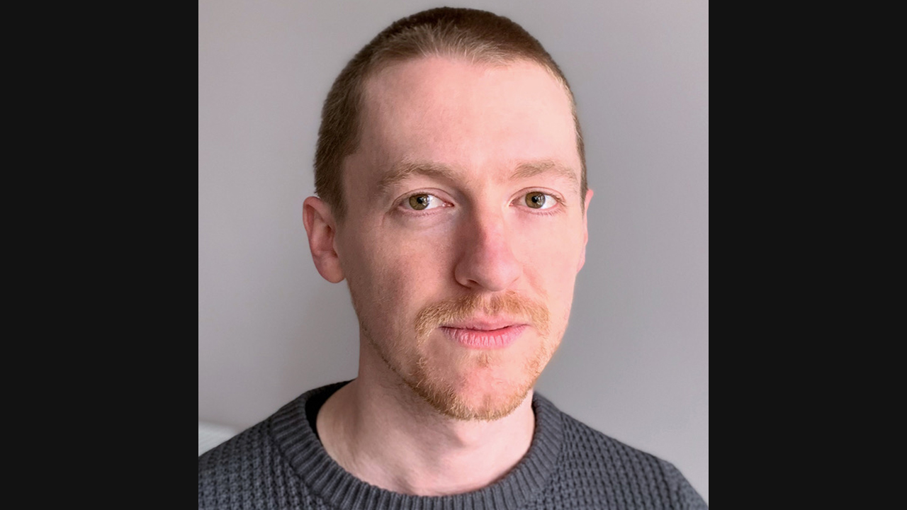 An image of a white male with shortly shaved hair. He is wearing a grey, wool jumper