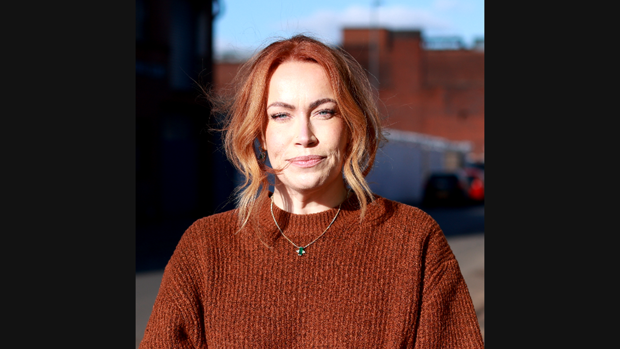 An image of a white woman with ginger hair, wearing a burnt orange jumper. 