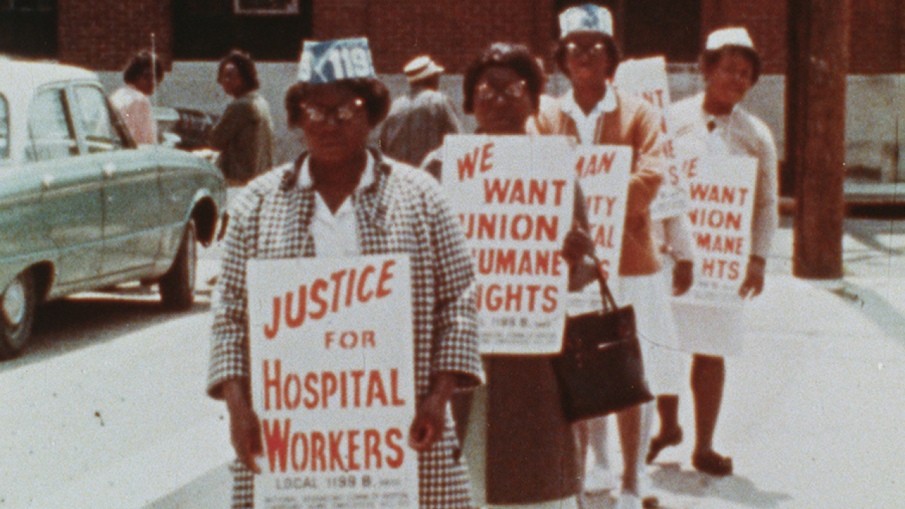 An archival image of four black, female nurses. They're stood in a line holding protest signs.