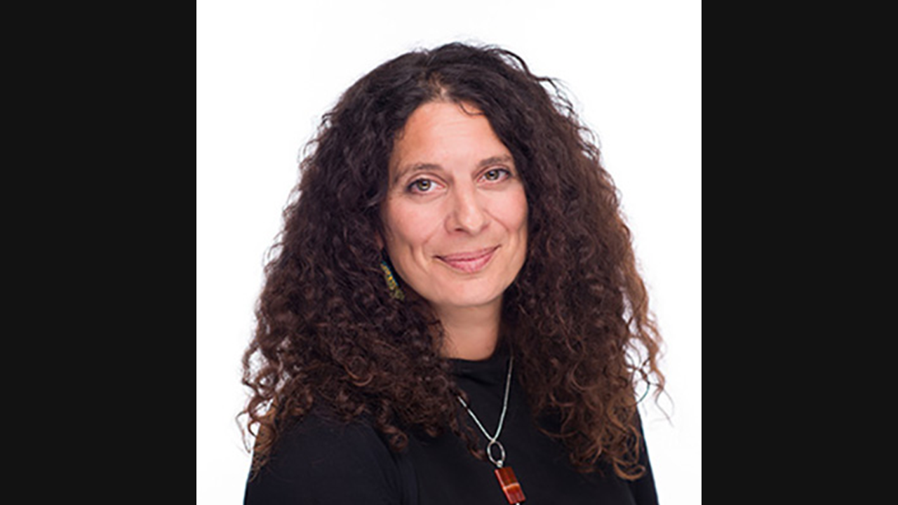 Headshot of a woman with dark brown, long, curly hair. She is wearing a black jumper and is pictured against a white backdrop.