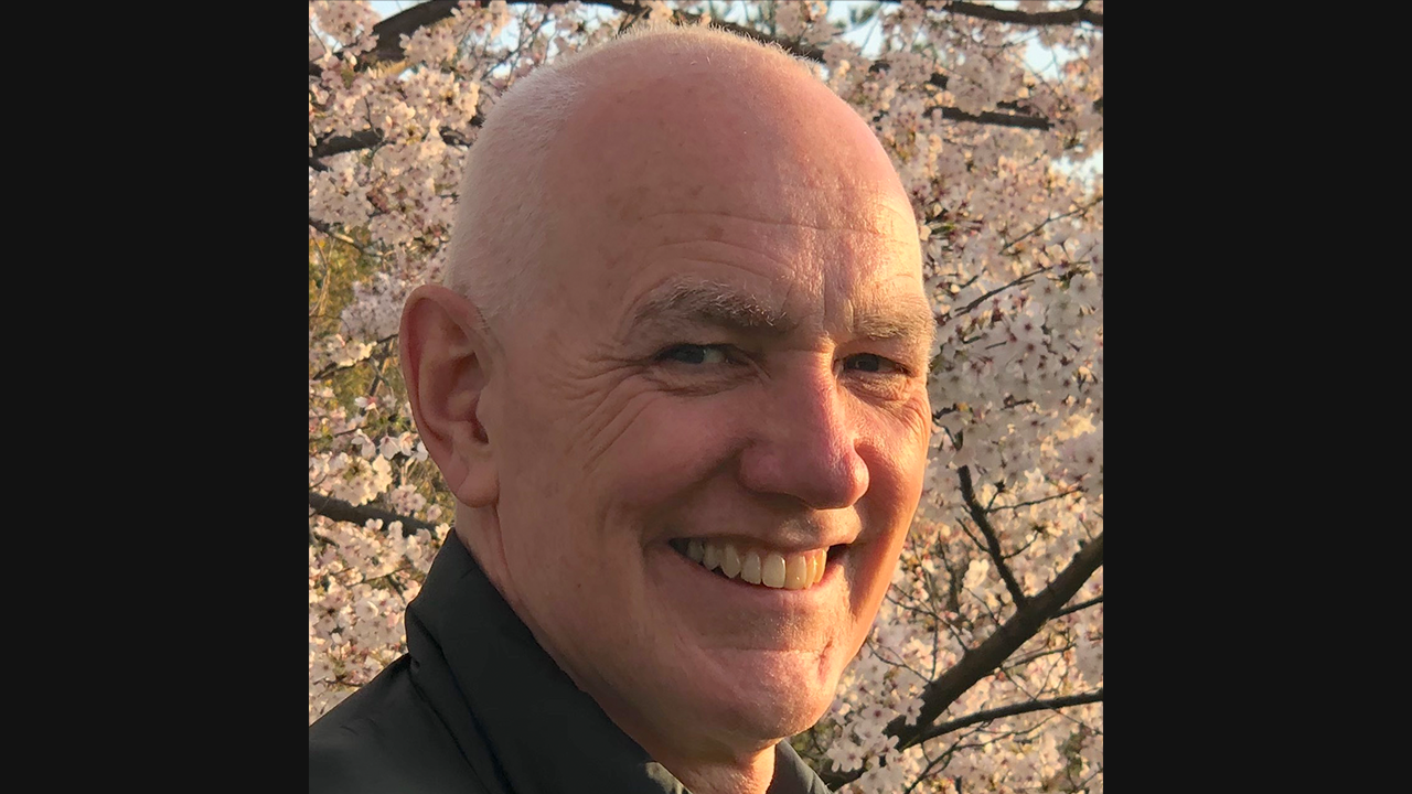 Image of an older, white male. He is smiling towards the camera, and is pictured against a blossom tree.
