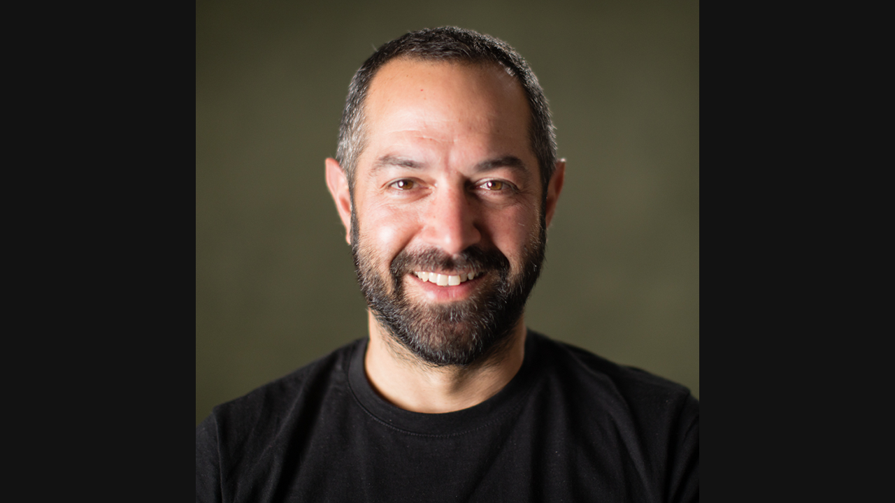 An image of a white male, with short dark hair and facial hair. He is wearing a dark t-shirt and is smiling towards the camera. He is pictured against a dark green background.