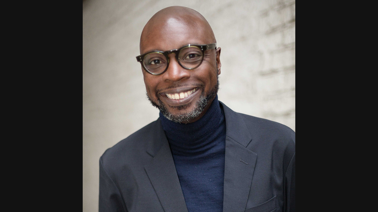 Image of a black, bald male with dark, greyish facial hair. He is wearing round framed glasses, a blue turtle neck and dark suit jacket. He is smiling towards the camera.