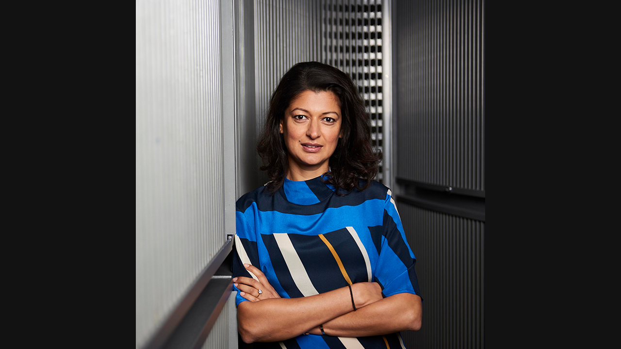 Image of a brown woman with long dark, straight hair. She is wearing a blue and white, geometric patterned blouse and has her arms folded across her chest. 