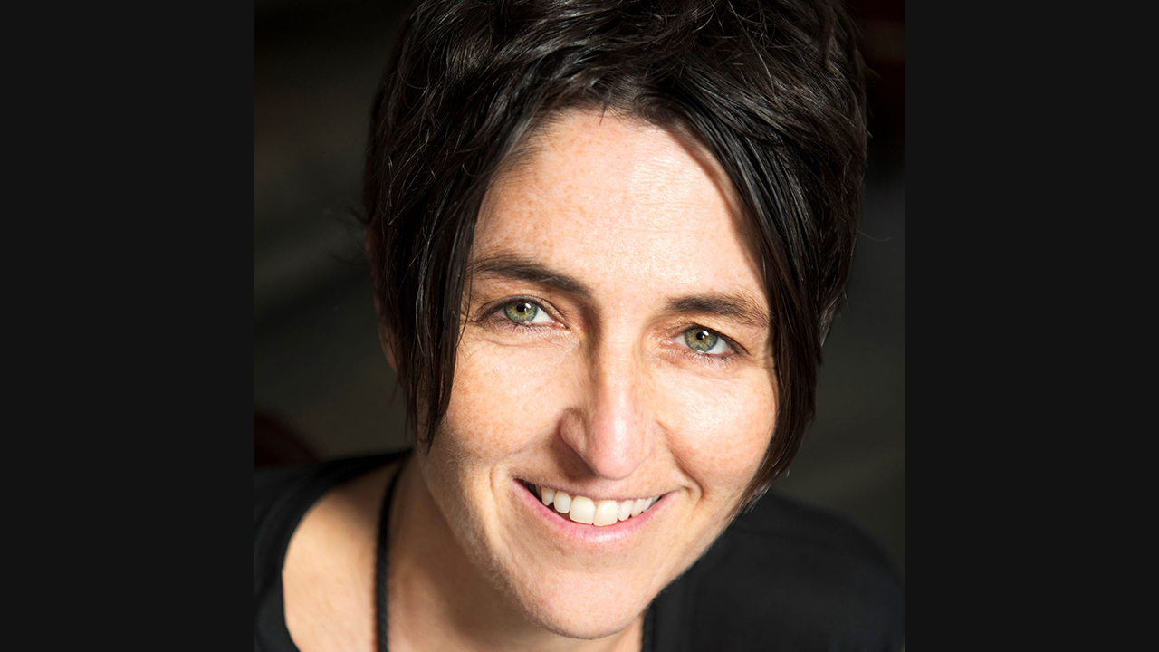 An image of a white woman with very short, dark brown hair. She's smiling towards the camera, and is pictured against a dark brown background.
