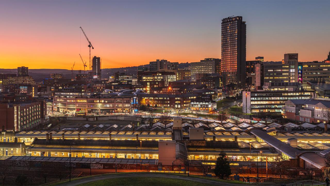 View of Sheffield skyline at dusk. Orange light can be seen from the windows of buildings. There are three cranes in the distance and the sky is turning a deep orange.