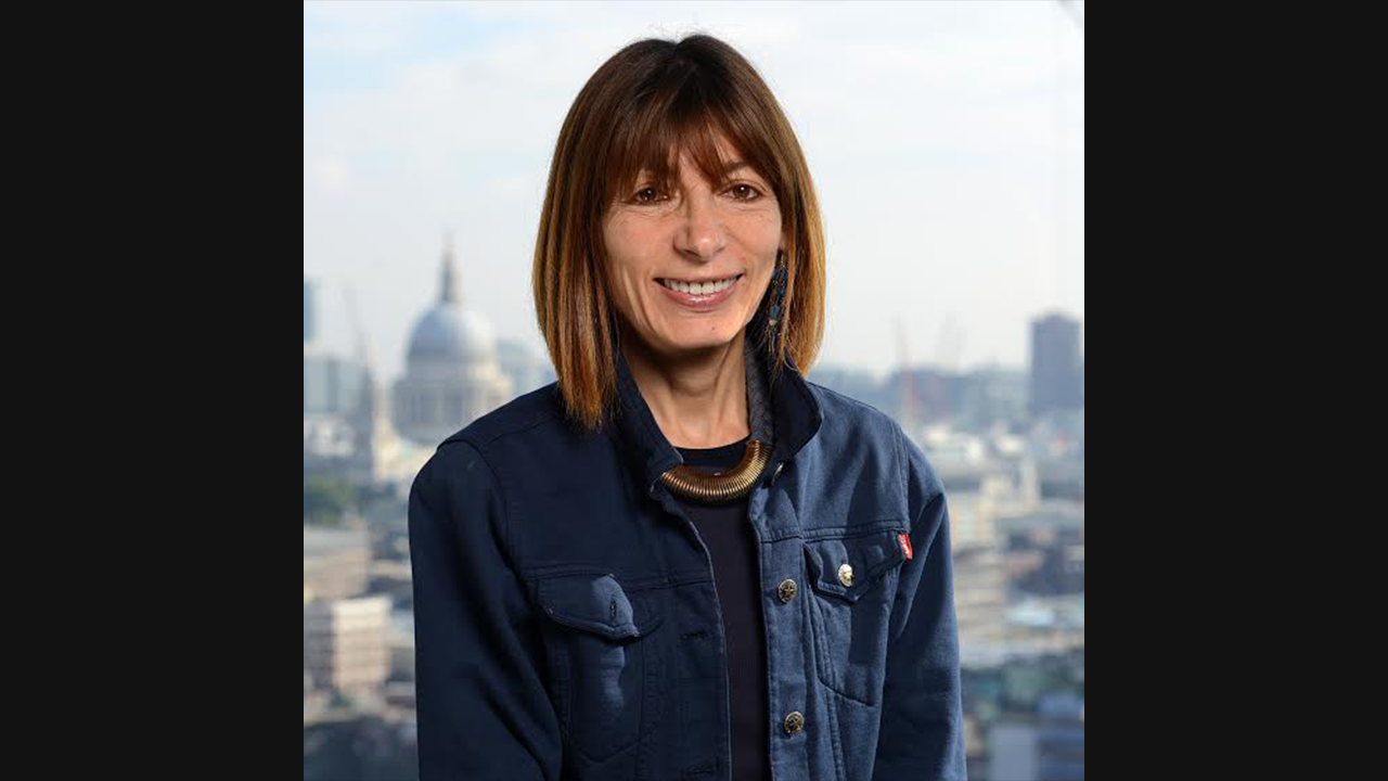 Image of an older, white woman with dark brown short hair and fringe. She is wearing a dark blue top and denim jacket, and is pictured against a city skyline.