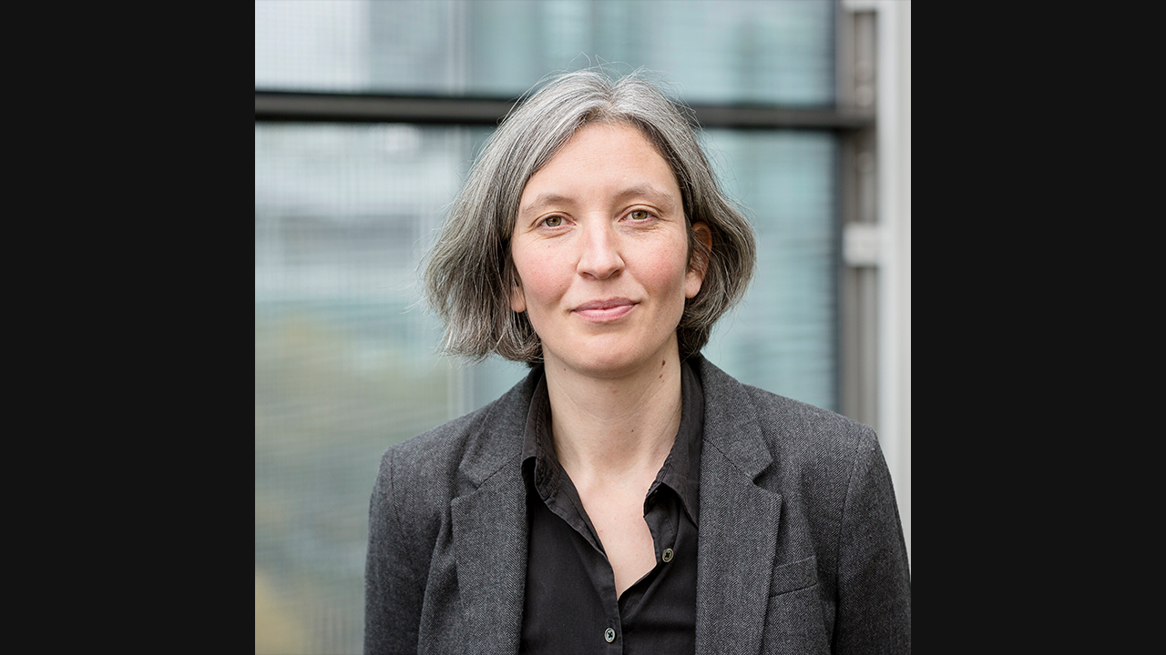 An image of a white woman, with short grey hair. She is wearing a dark shirt and grey suit jacket. 