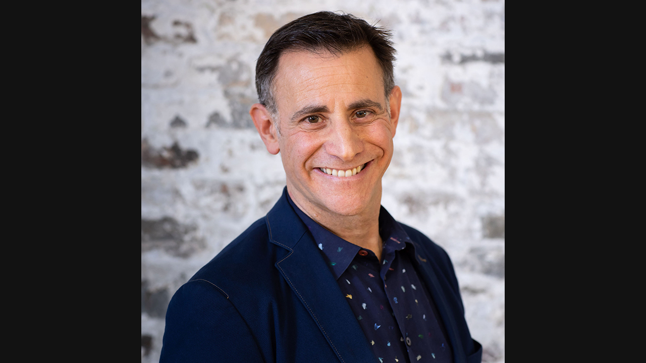 Image of a man with short dark hair, greying at the sides. He is smiling widely towards the camera. He is wearing a dark blue suit jacket, and dark blue shirt with multi-coloured dots. 