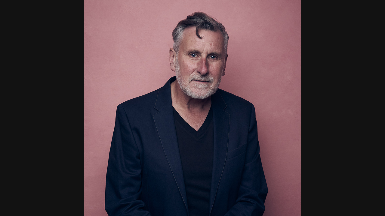 Image of a white, older male. He has dark grey hair, and facial hair. He's wearing a dark blue, v-neck top and suit jacket, and is pictured against a pink backdrop.