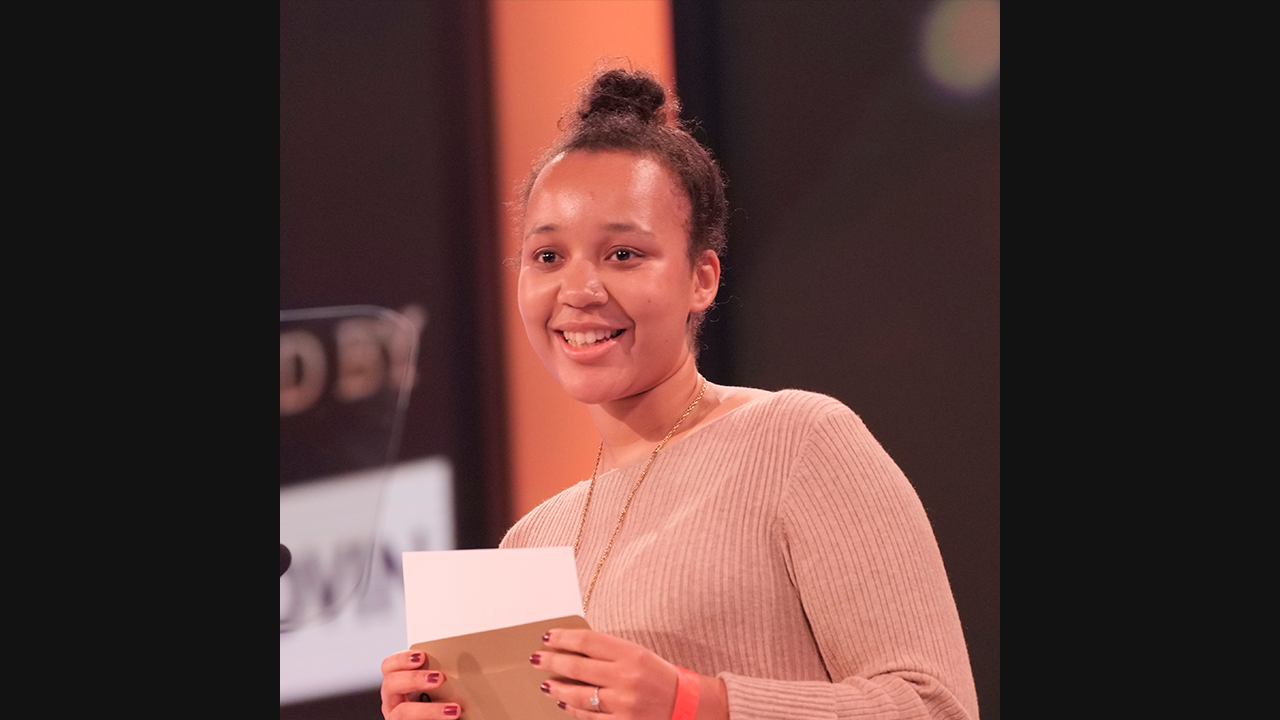 Image of a mixed-race woman, with dark hair tied up in a bun. She has a nose piercing, and is smiling. She is wearing a beige, ribbed top and is holding an envelope.