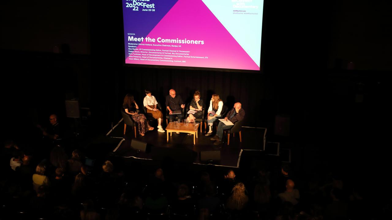 Aerial view of a full auditorium of people. A light is shining over a panel of speakers, behind them a screen reads: Meet the Commissioners