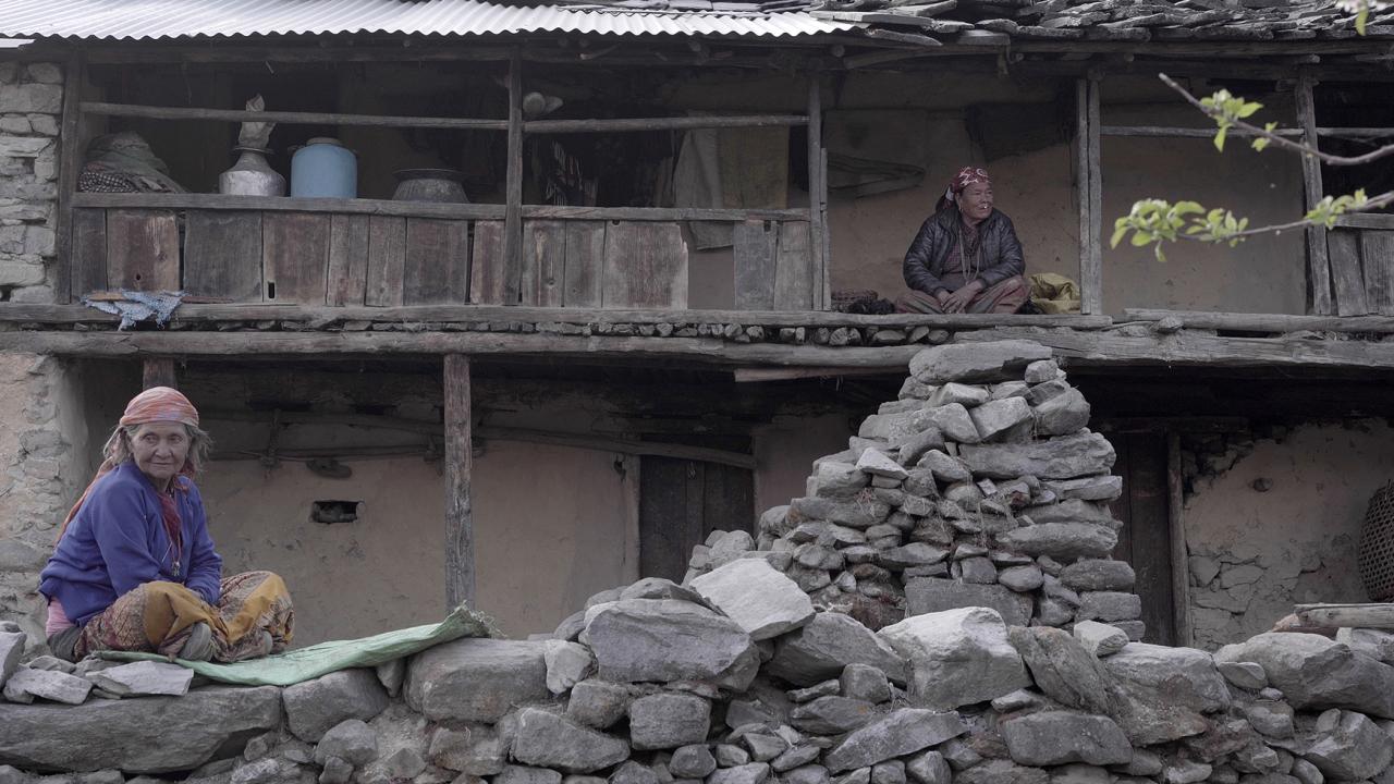 A woman sits on a stone wall. In the background, another woman sits on a balcony above a pile of rocks.