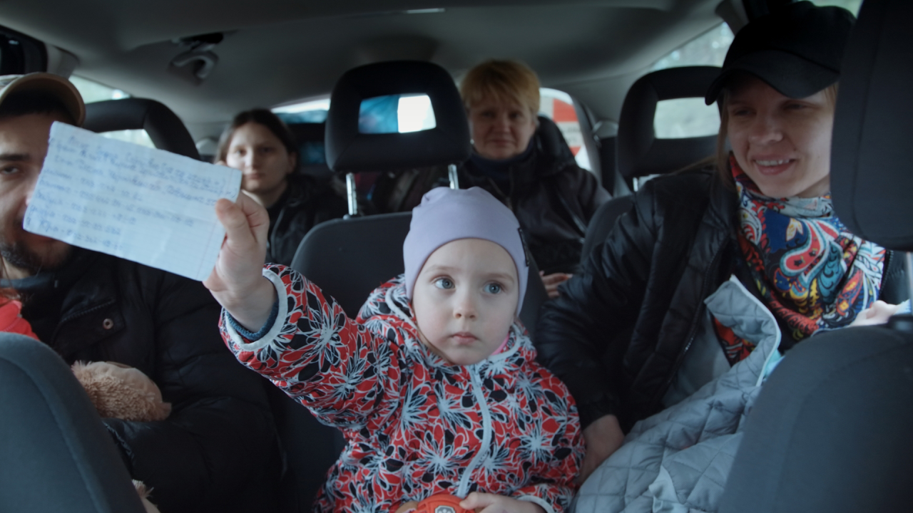 Image of a young child sat in a car. She is holding up a written sign on a piece of paper.