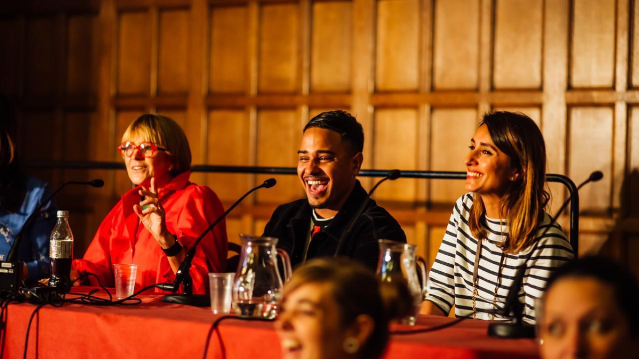 A panel of 3 people smiling whilst sat a table in front of microphones