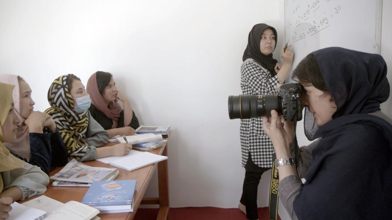 Women sat at a desk follow a teacher writing equations on the board. Another woman holds a camera.