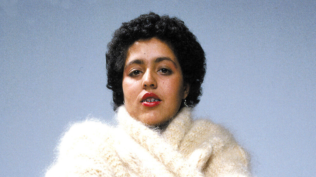 Image of Poly Styrene - a mixed-race woman with short, black curly hair. She is wearing red lipstick, and a white fluffy wool cardigan. She is pictured against a blue background.