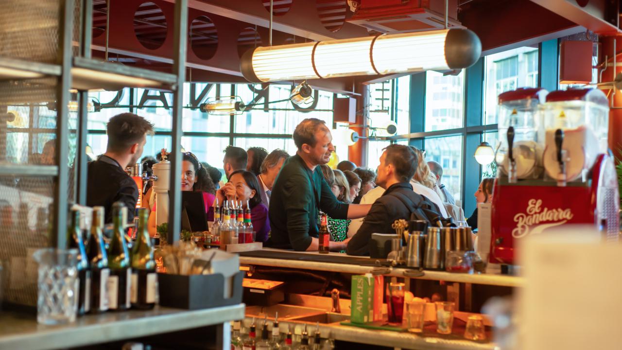Image from behind a bar. A crowd of people can be seen stood around the bar.
