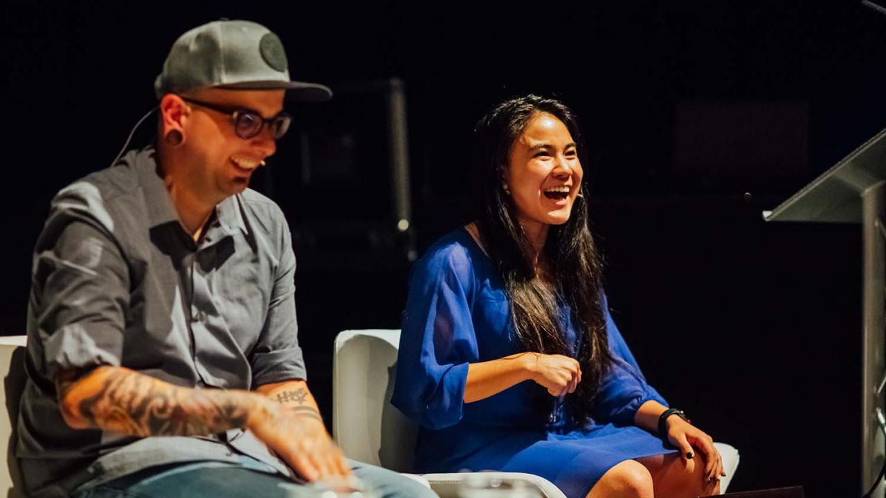 Image of a man and woman, sat in front of a lectern. They're both laughing. The man is wearing a grey shirt and cap. The woman is wearing a royal blue dress.