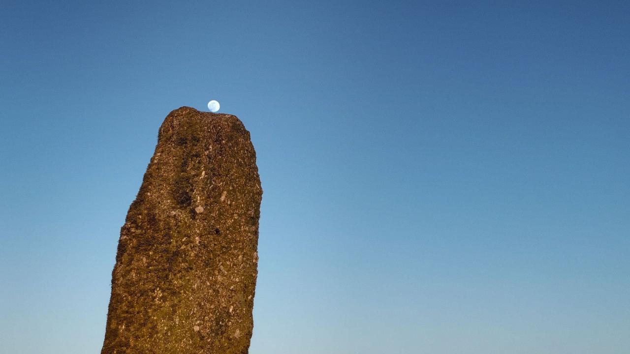 An image of a monolithic rock against a blue sky. The moon can be seen above the tip of the rock.
