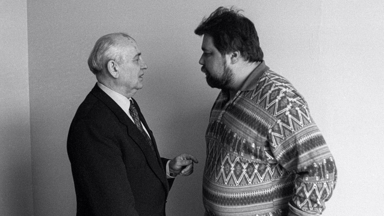 Black and white image of two men stood side-on, talking to each other. The man on the left is older and has sparse, grey hair. He's wearing a suit and pointing. The man on the right has a full head of dark hair, facial hair, and is wearing a patterned knit jumper. 