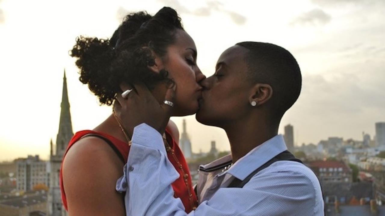 Image of two black women kissing, against a backdrop of a city skyline.