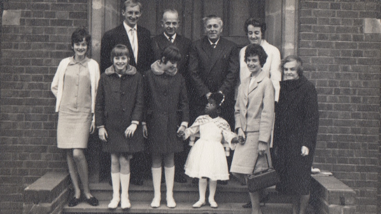 Black and white archival image. Nine white people pose for a photo with a black child. 