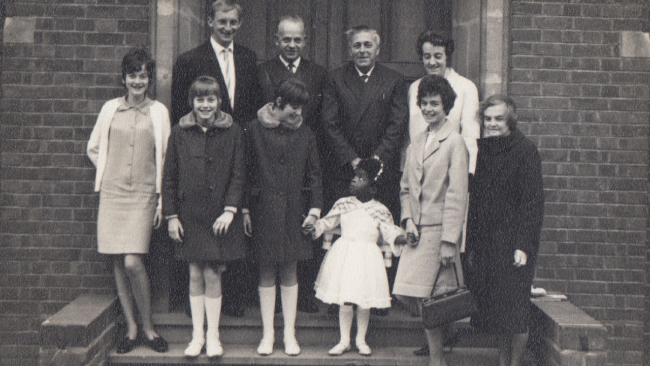 Black and white archival image. Nine white people pose for a photo with a black child.